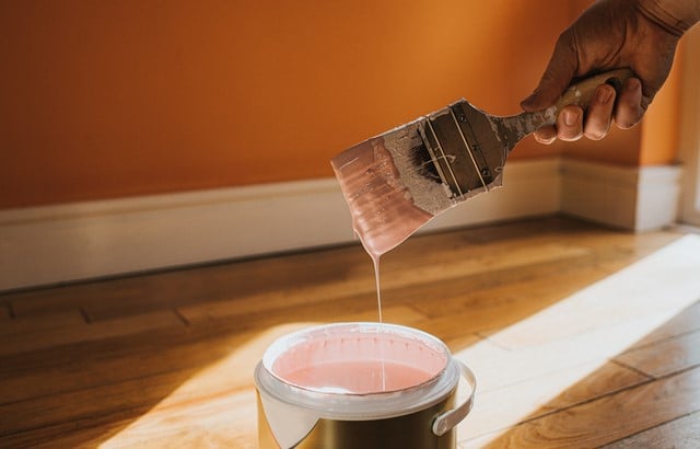 Hand dipping a paint brush into a large tin of pink paint as it drips back into the canister